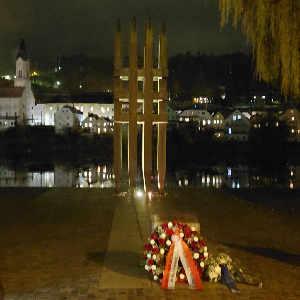 Das Mahnmal zum Gedenken der Opfer des Holocausts in Passau am Ufer des Inns bei Nacht. Im Hintergrund ist der Inn und die Innstadt mit erleuchteten Fenstern zu sehen.